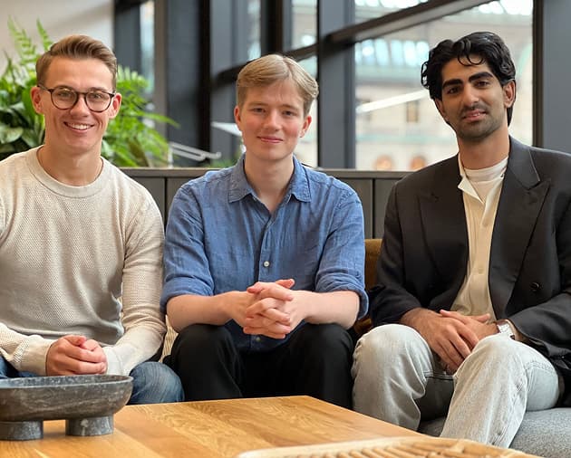 Three individuals sit closely together on a sofa, dressed in casual and smart attire, surrounded by greenery in a modern indoor space.